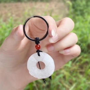 May include: A white jade keychain with a circular pendant. The pendant has a central hole and carved details. It is attached to a black cord, a red bead, and a silver keyring. The background is a blurred green.
