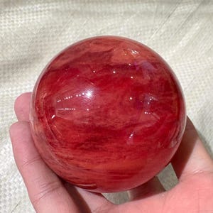 Pode incluir: Uma esfera vermelha translúcida polida, segurada em uma mão. A esfera tem um acabamento brilhante e um padrão de redemoinho de vermelho mais claro e branco. O objeto é um item decorativo, possivelmente uma bola de cristal ou um peso de papel.