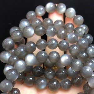 May include: A collection of grey moonstone beads. The spherical beads have a subtle sheen, with colour variations from light to dark grey. The beads are strung together, forming a necklace or bracelet.