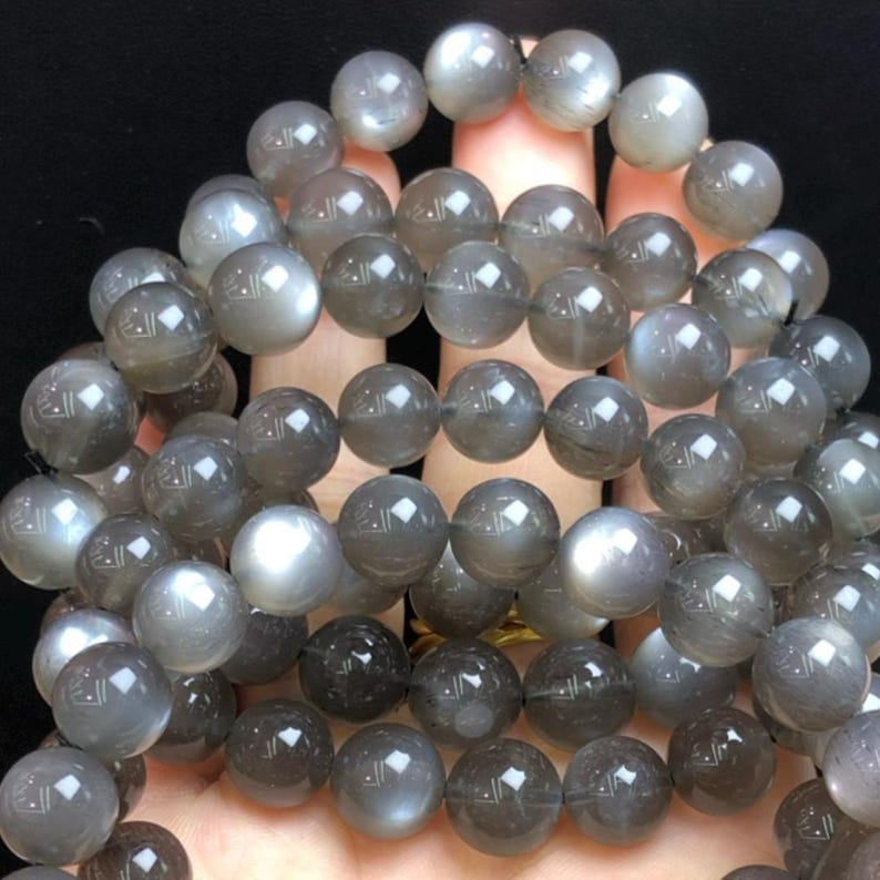 May include: A collection of grey moonstone beads. The spherical beads have a subtle sheen, with colour variations from light to dark grey. The beads are strung together, forming a necklace or bracelet.