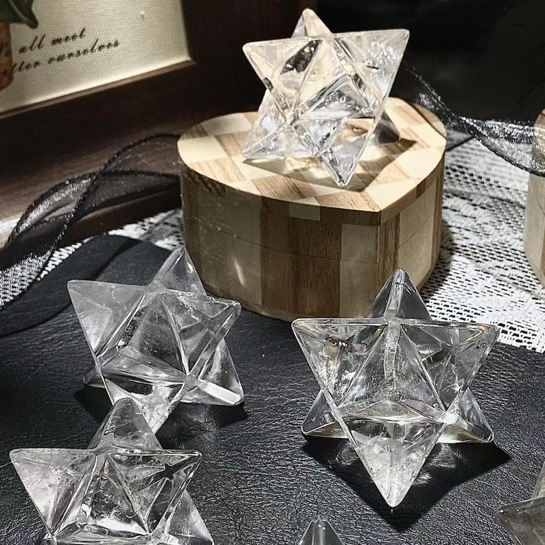May include: Clear crystal Merkaba star sculptures. The geometric shapes are displayed on a black surface and a wooden heart-shaped box. The Merkaba stars are transparent and faceted, reflecting light. The wooden box has a checkered pattern on the lid.