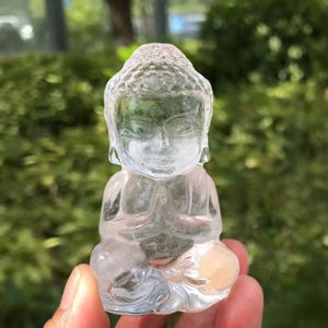 May include: A clear, translucent crystal Buddha statue in a seated position with hands clasped in prayer. The statue has detailed facial features and is held in a hand against a blurred green background.