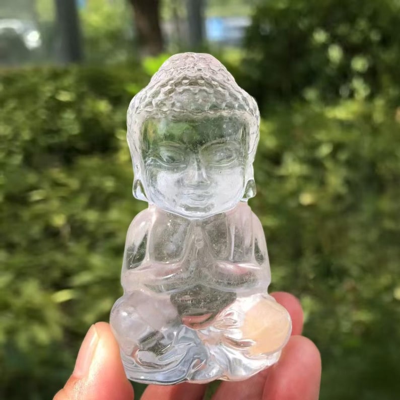 May include: A clear, translucent crystal Buddha statue in a seated position with hands clasped in prayer. The statue has detailed facial features and is held in a hand against a blurred green background.