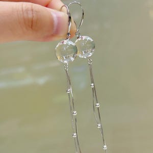 以下が含まれることがあります： シルバーのドロップピアス。各イヤリングは、上部に透明な球形のビーズがあり、細い銀色のチェーンと小さなビーズが均等に配置されています。フック留めです。