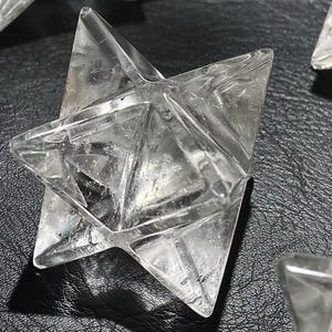 May include: Clear quartz Merkaba star, a geometric crystal with six points. The star is transparent, allowing light to pass through, and is displayed on a dark surface. The Merkaba star is a spiritual and metaphysical tool.