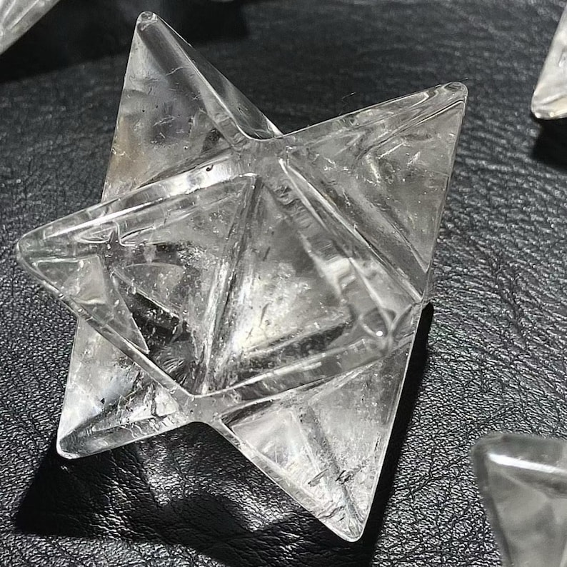 May include: Clear quartz Merkaba star, a geometric crystal with six points. The star is transparent, allowing light to pass through, and is displayed on a dark surface. The Merkaba star is a spiritual and metaphysical tool.