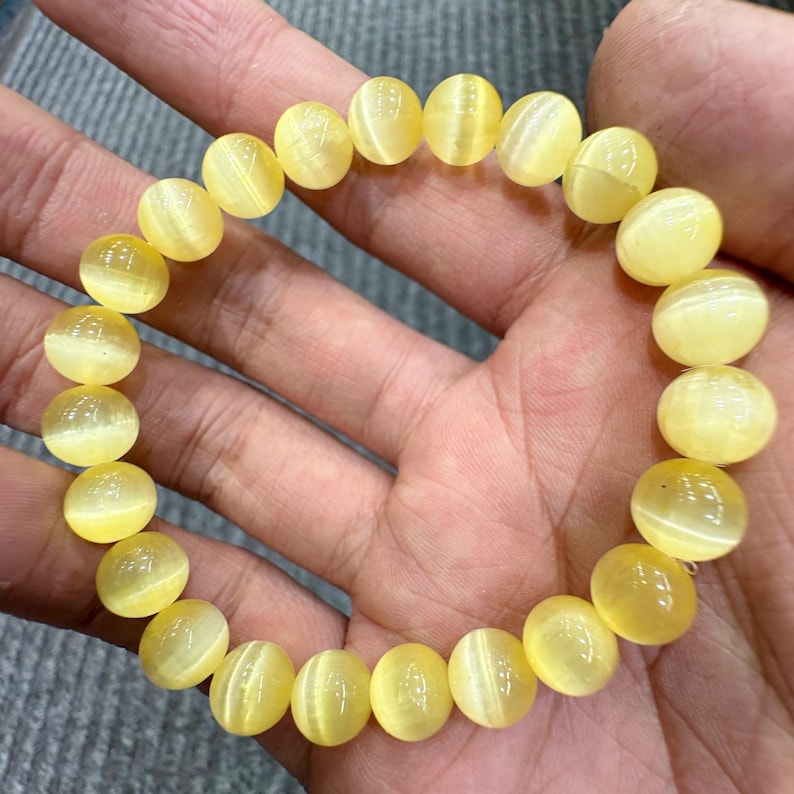 May include: A yellow cat's eye gemstone bracelet. The bracelet is made of round, polished beads with a chatoyant effect. The beads are a light yellow color and are strung together on an elastic band.