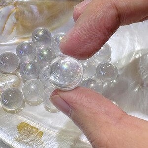 May include: A collection of small, spherical, clear glass beads with an iridescent finish that reflects rainbow colors. Each bead has a smooth, polished surface and a translucent quality. The beads are grouped together, showing their reflective properties.
