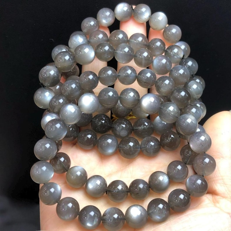 May include: Multiple strands of grey moonstone bead bracelets. The round beads have a subtle sheen, with colour variations from light grey to darker shades. The bracelets are shown against a dark background.
