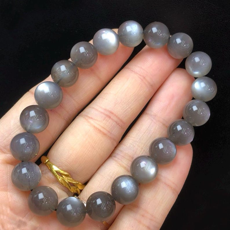 May include: A bracelet of grey moonstone beads, each approximately 1 cm in diameter. The beads have a soft sheen and are strung on a black cord. The bracelet is held in a hand, highlighting the natural beauty of the stones.