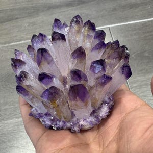 Könnte beinhalten: Ein Cluster aus Amethystkristallen, mit tiefvioletten Spitzen, die in eine blasse, durchscheinende Basis übergehen. Die Kristalle sind dicht gepackt und bilden einen strukturierten, natürlichen Cluster. Die Gesamtform ist abgerundet und kompakt.