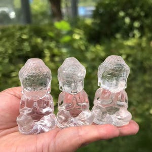 May include: Three clear quartz Buddha figurines in a seated meditation pose. The translucent statues have detailed facial features and are held in a hand against a blurred green background. The figurines are approximately 2 inches tall.