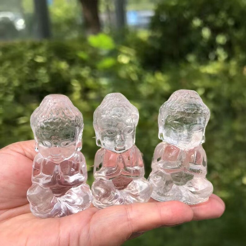 May include: Three clear quartz Buddha figurines in a seated meditation pose. The translucent statues have detailed facial features and are held in a hand against a blurred green background. The figurines are approximately 2 inches tall.
