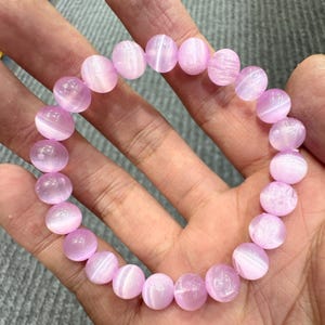 May include: A bracelet featuring round, light purple beads. The beads have a translucent quality with subtle white striations. The bracelet is held in a hand, showcasing the delicate color and texture of the gemstones. The beads are approximately 0.5 inches in diameter.