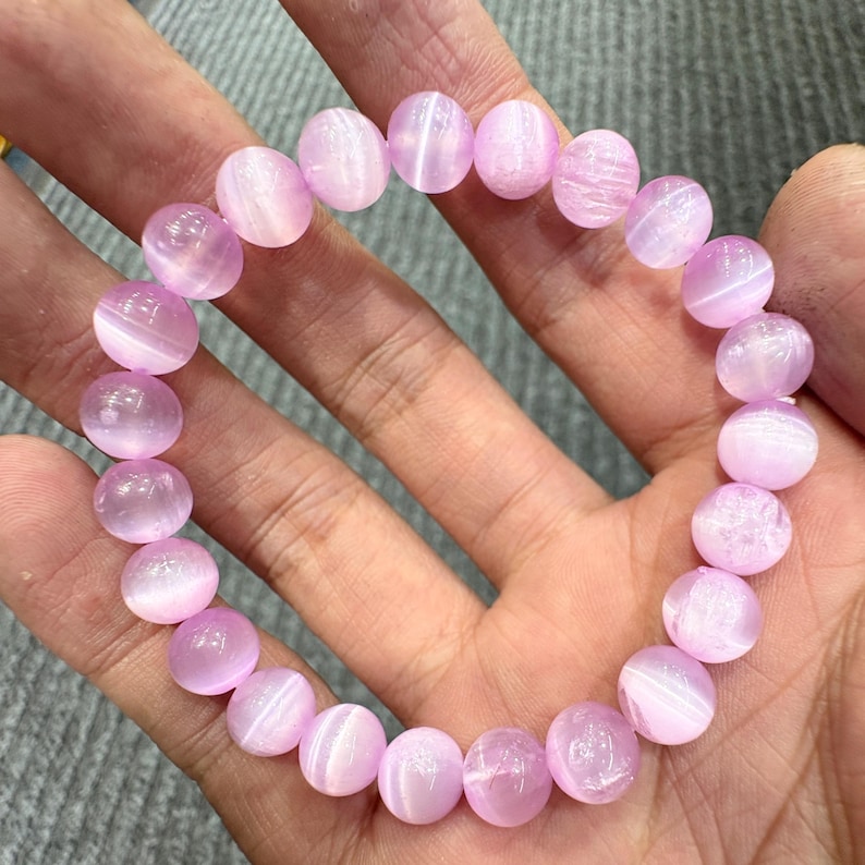 May include: A bracelet featuring round, light purple beads. The beads have a translucent quality with subtle white striations. The bracelet is held in a hand, showcasing the delicate color and texture of the gemstones. The beads are approximately 0.5 inches in diameter.