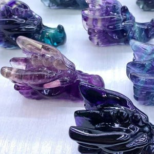 May include: A selection of carved dragon figurines in shades of purple, blue, and clear fluorite crystal. The dragons are detailed with visible wings and scales, and are displayed on a white surface.