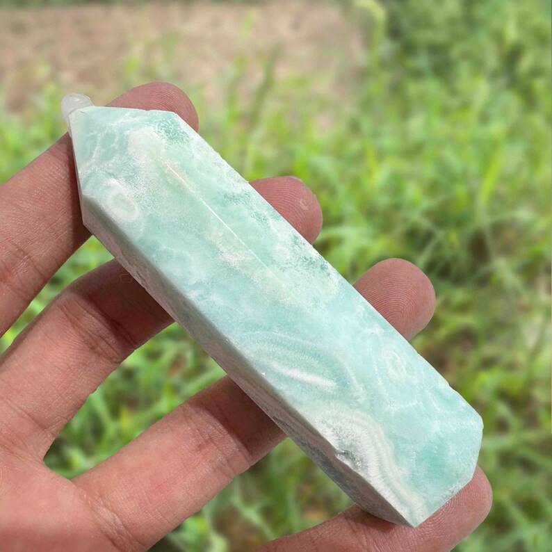 May include: A light blue crystal point with white marbling. The polished stone is held in a hand, with a blurred green background. The crystal is a rectangular prism shape, with a pointed top. The crystal is approximately 5 inches long.