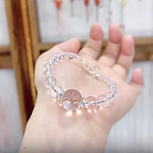 May include: A clear quartz crystal bracelet with a large, round, pinkish-brown central bead. The bracelet features smaller, clear beads strung together, creating a delicate accessory. The bracelet is held in a hand.