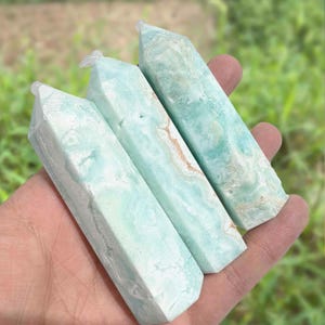May include: Three polished, rectangular crystals with a light blue and white marbled appearance are held in a hand. The crystals have a pointed top and a faceted design. The background is blurred green.