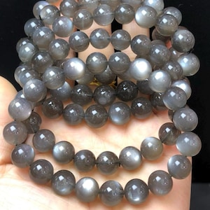 May include: Multiple strands of grey moonstone bead bracelets. The round beads have a subtle sheen, with colour variations from light grey to darker shades. The bracelets are displayed on a hand.