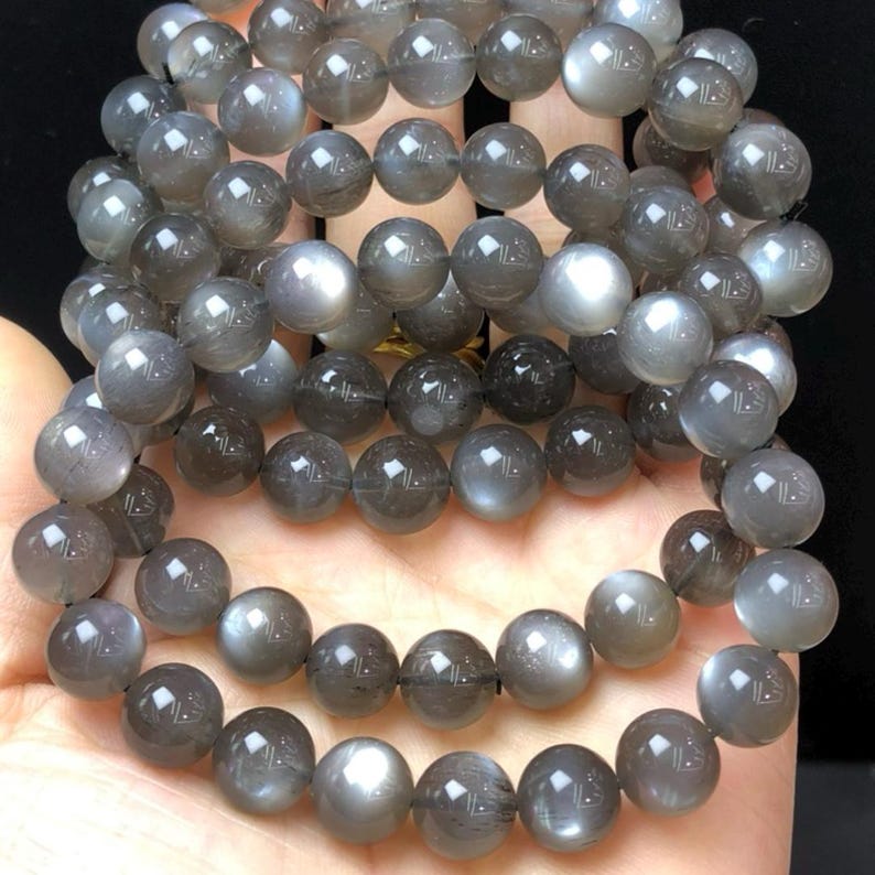 May include: Multiple strands of grey moonstone bead bracelets. The round beads have a subtle sheen, with colour variations from light grey to darker shades. The bracelets are displayed on a hand.
