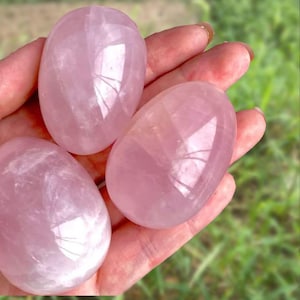 Op de afbeelding: Drie gepolijste rozenkwarts stenen in een ovale vorm. De stenen hebben een zachte roze kleur en een glad, reflecterend oppervlak. De stenen worden in een hand gehouden, met een vage groene achtergrond.