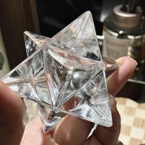 May include: A clear, multi-faceted crystal star, held in a hand. The star has a geometric design with sharp angles and a transparent appearance. The crystal reflects light, creating a sparkling effect. The object is likely a decorative item or a spiritual tool.