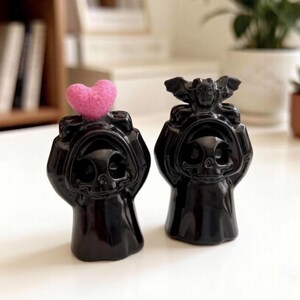 May include: Two black ceramic figurines shaped like hooded figures with skull-like faces. The left figure holds a textured pink heart above its head, while the right figure holds a small black bat with outstretched wings above its head.