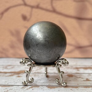 Può includere: Una sfera grigio scuro con una superficie strutturata, esposta su un supporto decorativo in metallo argentato. Il supporto ha tre gambe ornate e una parte superiore circolare, su uno sfondo sfocato.
