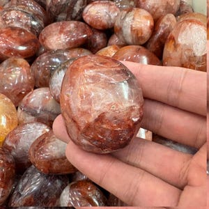 Può includere: Una mano tiene in mano un cristallo di quarzo rosso lucido, di forma ovale. Il cristallo presenta venature e inclusioni interne, con un aspetto traslucido. Altri cristalli simili sono sullo sfondo, creando una collezione di pietre preziose naturali.