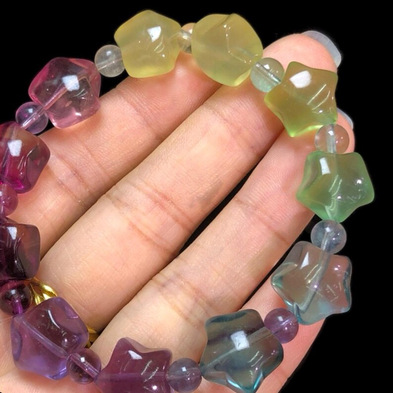 May include: A colorful bracelet featuring star-shaped and faceted beads in shades of purple, pink, green, and yellow. The bracelet is strung with smaller, round beads and has a gold clasp. The gemstones are translucent, and the bracelet is displayed against a black background.