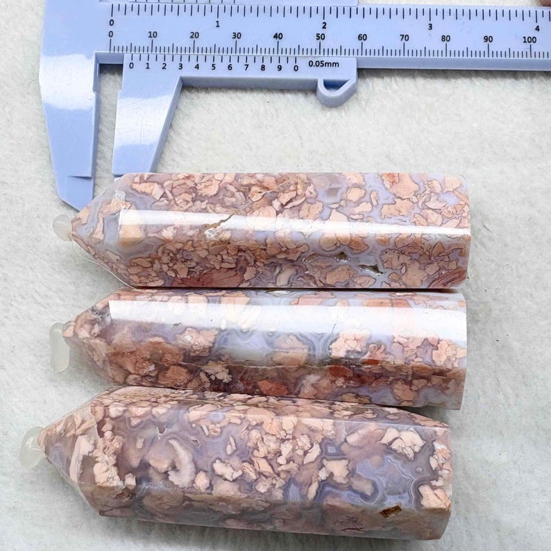 May include: Three polished gemstone obelisks with a patterned surface in shades of brown, beige, and gray. A blue measuring tool is in the background, showing measurements in millimeters.