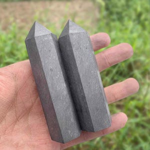 Puede incluir: Dos obeliscos de cristal gris facetados se sostienen en una mano sobre un fondo verde borroso. Los obeliscos tienen forma rectangular con una parte superior puntiaguda. Los cristales son de color gris oscuro sólido.