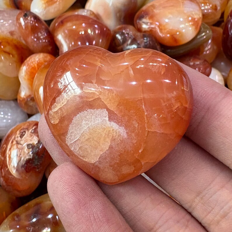 May include: A heart-shaped, polished carnelian stone with a warm orange hue and white inclusions. The stone is held in a hand, with other polished stones in the background. The heart is approximately 2 inches wide.