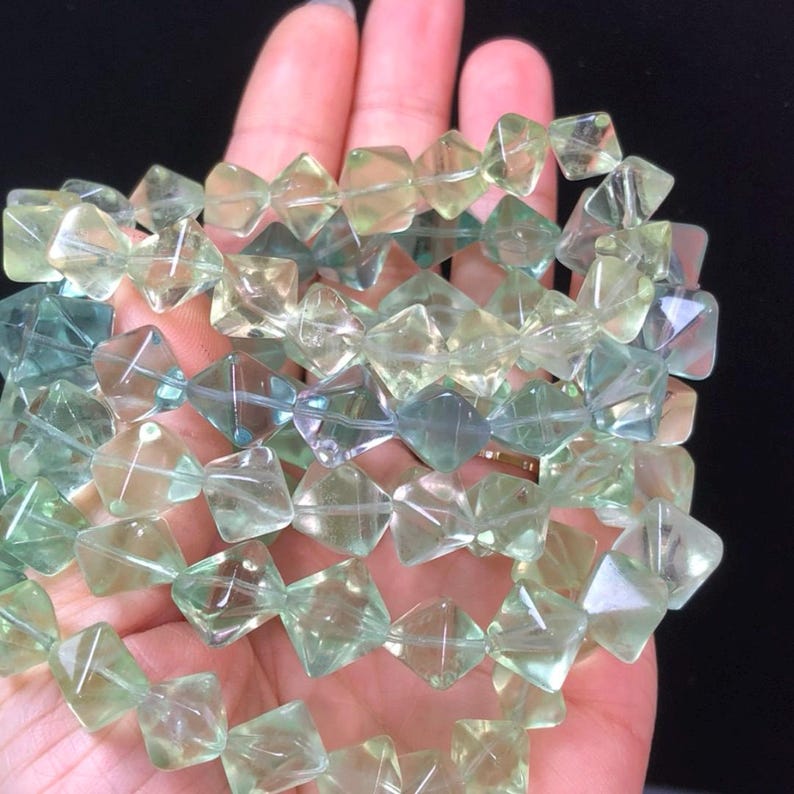 May include: A strand of faceted, translucent beads in shades of green and yellow. The beads are square-shaped with a diamond-like cut, creating a sparkling effect. The beads are held in a hand, showcasing their color and clarity.