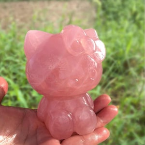 Peut inclure: Figurine rose translucide d'un chat de dessin animé avec un nœud. Le chat est en position assise et est fait d'un matériau lisse et poli. La figurine est tenue dans une main sur un fond vert flou.