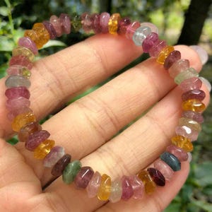 May include: A multi-colored gemstone bracelet. The bracelet features a variety of small, irregularly shaped stones in shades of pink, yellow, orange, green, and blue. The stones are strung together to form a flexible band.