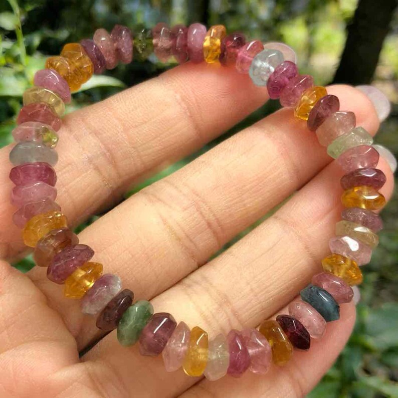 May include: A multi-colored gemstone bracelet. The bracelet features a variety of small, irregularly shaped stones in shades of pink, yellow, orange, green, and blue. The stones are strung together to form a flexible band.