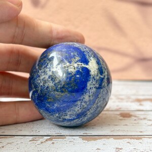 以下が含まれることがあります： 研磨された、球形のラピスラズリの宝石。濃い青色の石は、白と金色の斑点模様です。滑らかで反射する表面を強調するように、球は手で持たれています。天然の装飾品です。