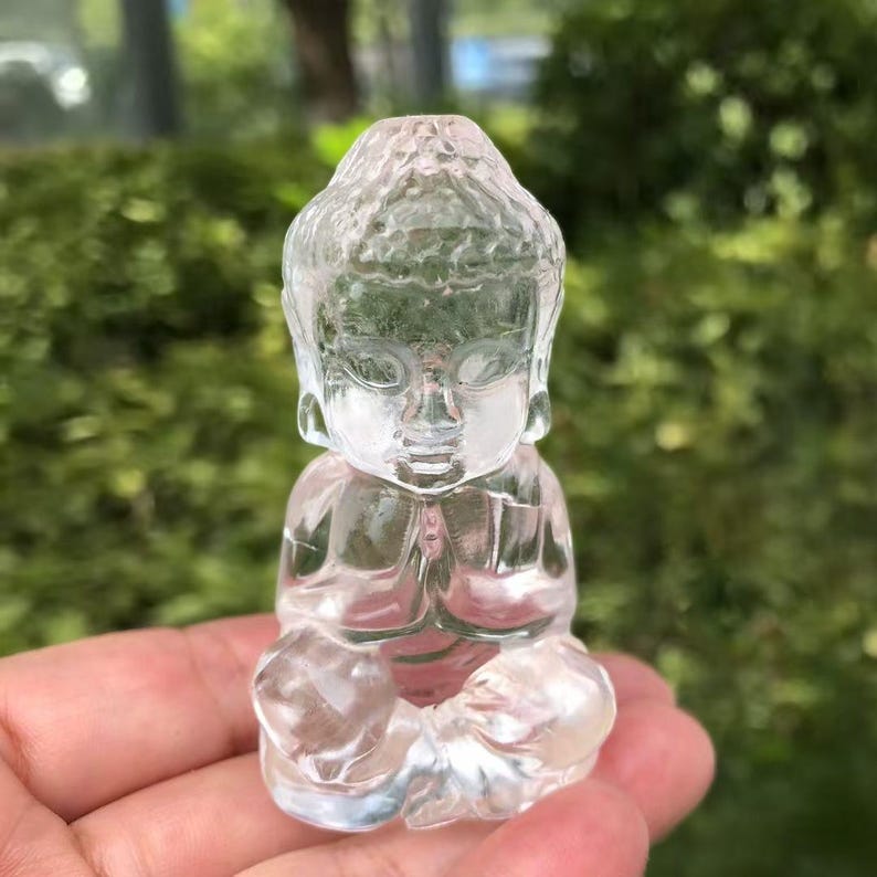 May include: A clear, transparent crystal Buddha statue. The Buddha is depicted in a seated position with hands clasped in prayer. The statue has detailed facial features and is held in a hand against a blurred green background.