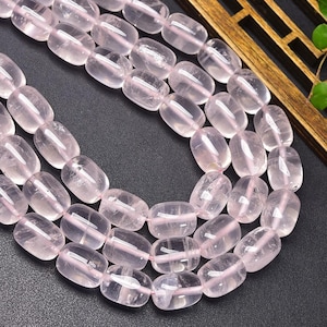 Puede incluir: Primer plano de hileras de cuentas de cuarzo rosa ovaladas y pulidas. Las cuentas son de color rosa pálido y están ensartadas, formando un collar o pulsera. Las cuentas son translúcidas.
