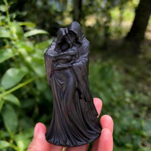 Puede incluir: Una escultura negra que representa dos figuras encapuchadas abrazándose. Las figuras tienen túnicas y capuchas detalladas, creando una estética gótica o fantástica. La escultura se sostiene en una mano sobre un fondo verde borroso.