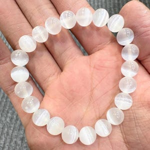 May include: A white beaded bracelet composed of round, translucent beads. The bracelet is made of polished, spherical beads strung together. The beads have a subtle, internal pattern. The bracelet is shown resting on a hand.