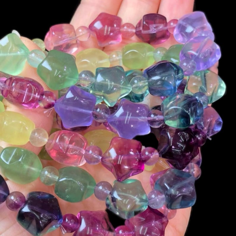 May include: A close-up of a collection of colorful gemstone beads. The beads are in various shapes, including stars and rounded forms, and in shades of pink, purple, green, yellow, and blue. The beads are strung together, creating a vibrant and eye-catching accessory.