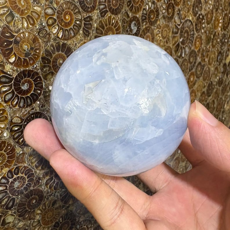 May include: A light blue sphere made of a crystalline material. The sphere is held in a hand, and the background features a pattern of brown and tan spiral shapes. The sphere appears to be approximately 3 inches in diameter.