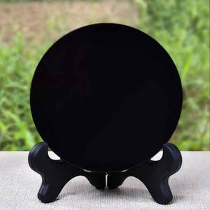 K&ouml;nnte beinhalten: Eine schwarze, runde Dekoplatte auf einem schwarzen, dreibeinigen St&auml;nder. Die Platte hat eine gl&auml;nzende Oberfl&auml;che und ist mittig platziert. Der St&auml;nder besteht aus dunklem Material und steht auf einer hellen Oberfl&auml;che, mit verschwommenem gr&uuml;nem Hintergrund.