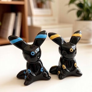 Könnte beinhalten: Zwei glänzende schwarze Keramikfiguren in Form von Wesen mit spitzen Ohren. Eine Figur hat leuchtend blaue Ringmarkierungen an Ohren, Stirn und Gliedmaßen. Die andere Figur zeigt identische Ringmarkierungen in einer kräftigen gelben Farbe.