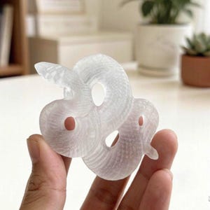 Peut inclure: Une figurine en cristal sculpté, translucide et transparente, en forme de serpent enroulé. La sculpture présente un motif d'écailles texturé et détaillé sur toute sa surface, une tête lisse et deux découpes circulaires dans le corps du serpent.