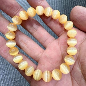 May include: A golden yellow beaded bracelet. The bracelet is made of round, polished beads with a subtle striped pattern. The beads are strung together to form a complete circle. The bracelet is held in a hand.