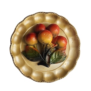 May include: A decorative, round, cream-colored plate with a raised relief of fruit. The fruit is a mix of red and yellow, with green leaves. The plate has a scalloped edge with a gold rope-like trim.